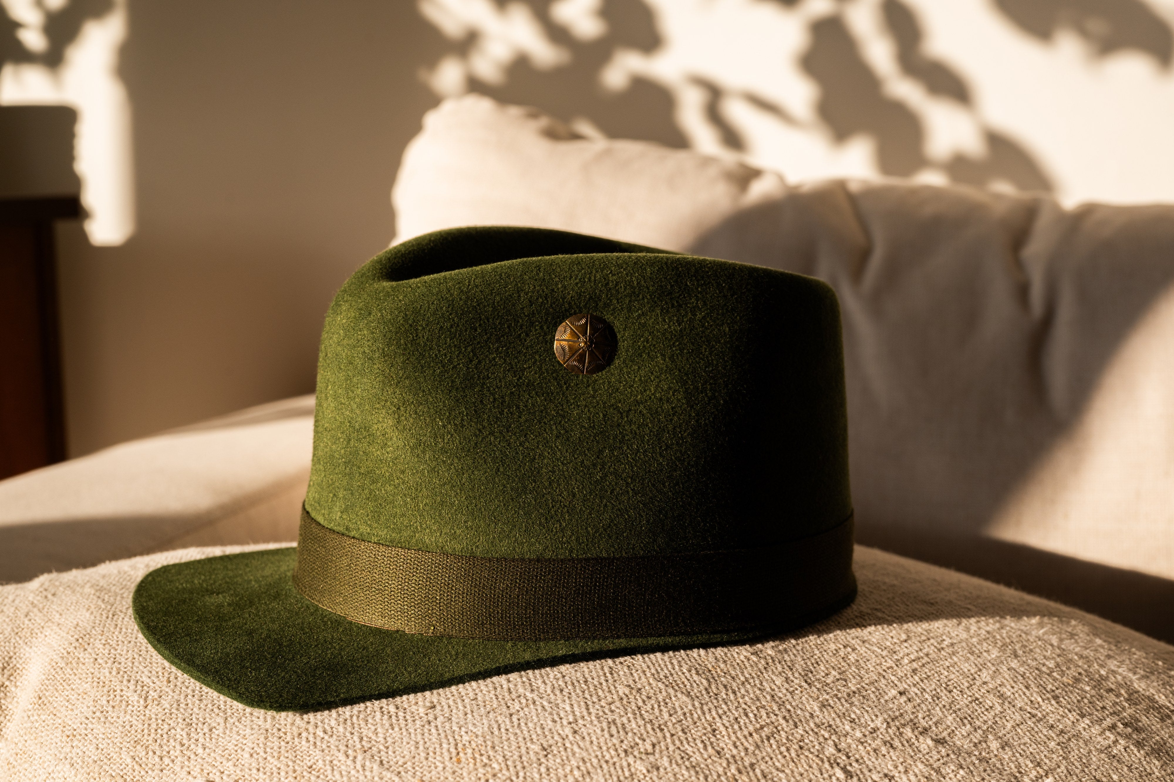 Hilltop olive green hat with brass stud, side view on linen in dappled sunlight.